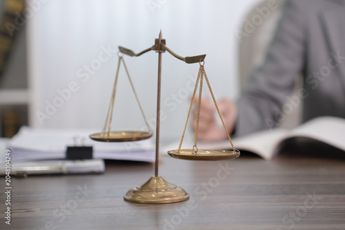 Fotografie A pair of vintage golden scales of justice rests on a wooden desk with a blurred figure of a lawyer and a book in the background