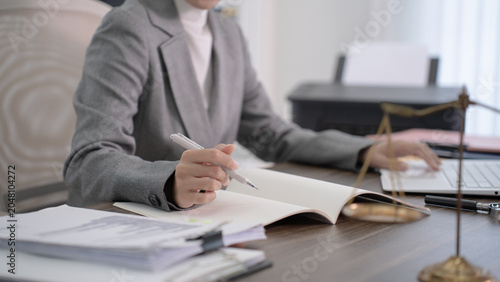 A professional woman in a grey suit sits at a desk and holds a pen while working on paperwork with a laptop nearby.