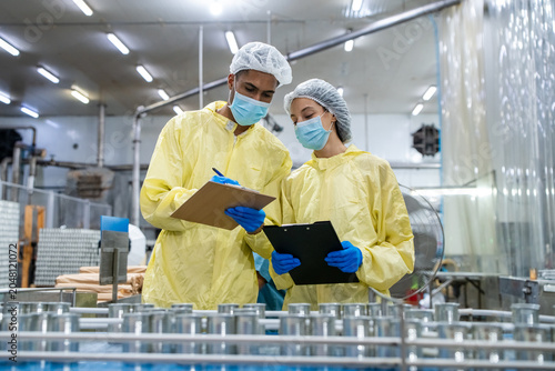 Two workers wear yellow lab coats, hairnets, masks, and gloves. They hold clipboards and review documents. Metal shelves and machinery surround the facility