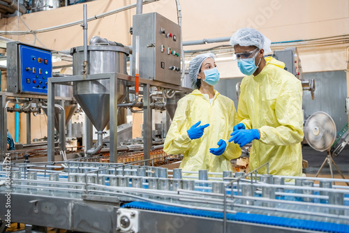 Two workers wear yellow protective suits and blue gloves. They have hairnets and face masks on. Metal conveyors and control panels are visible in the background