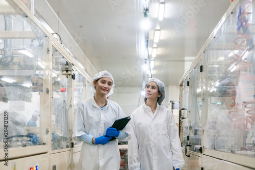 Two women in white lab coats and hairnets stand in a cleanroom. One holds a tablet with blue gloves. Glass partitions separate lab sections. Bright lights illuminate the sterile space
