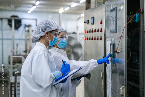 Two individuals wear white lab coats, hairnets, and face masks. One points to a machine while the other holds a clipboard. They use blue gloves during a technical check. The environment is a clean
