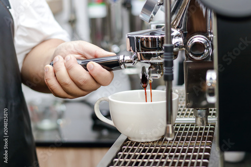 A professional barista making fresh espresso, holding a portafilter handle while hot coffee flows into a white ceramic cup from a modern espresso machine in a cafe.