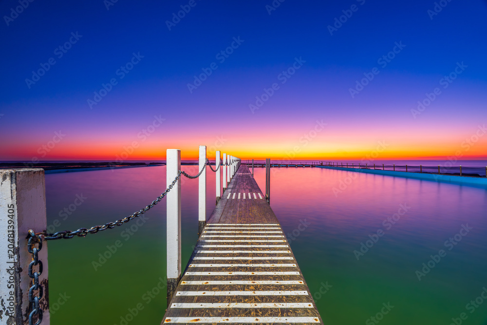 Fototapeta premium Leading lines from the timber swimming platform guide the eye across North Narrabeen Rockpool at sunrise, featuring calm water, pastel skies and Sydney coastal scenery.
