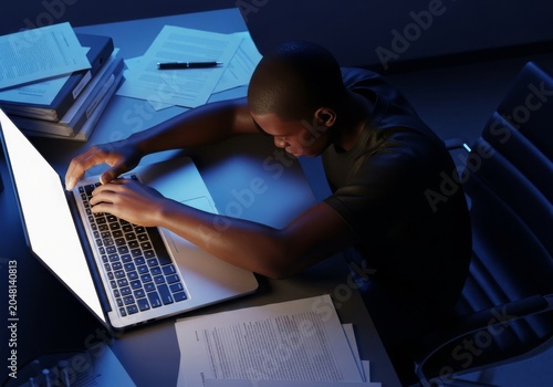 A dimly lit office desk with scattered files and a laptop, where dark-skinned arms rest heavily, portraying a mood of deep hopelessness, desk, technology, anxiety