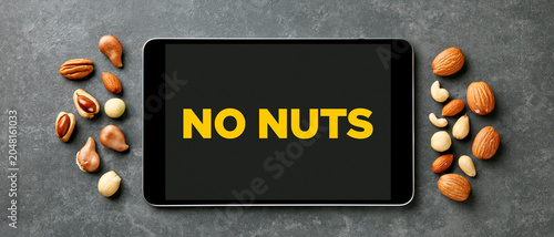 A smartphone displaying the text NO NUTS surrounded by various nuts on a dark, textured surface. The image emphasizes a prohibition or warning related to nuts.