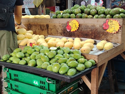 Customer browsing a vibrant display of organic raw mangoes and fresh avocados at a traditional produce market.