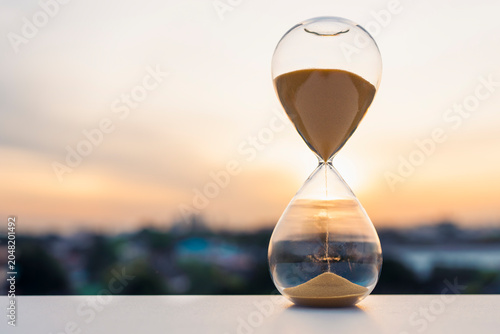 Schilderij op canvas Hourglass with falling sand on table against blurred sunset city background