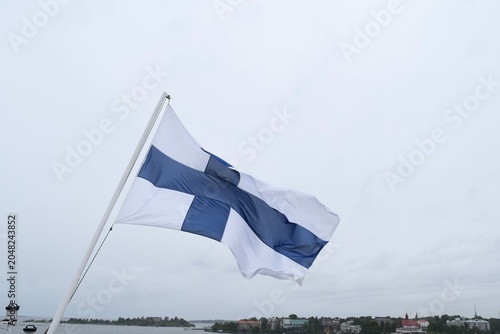 National flag of Finland waving on a flagpole against a cloudy sky with Helsinki coast in the background 