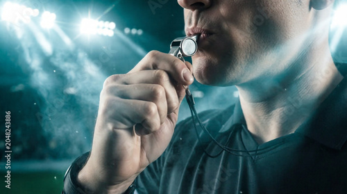 Close-up of referee blowing silver whistle at packed floodlit stadium.
