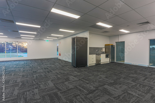 Tableau sur toile Commercial office interior featuring open workspace, kitchenette facilities, carpet tiles, large windows and modern ceiling lighting in a professional business environment