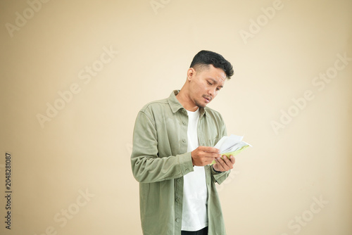 Foto Stressed young Asian man in olive green shirt counting cash with worried expression symbolizing financial pressure and economic crisis