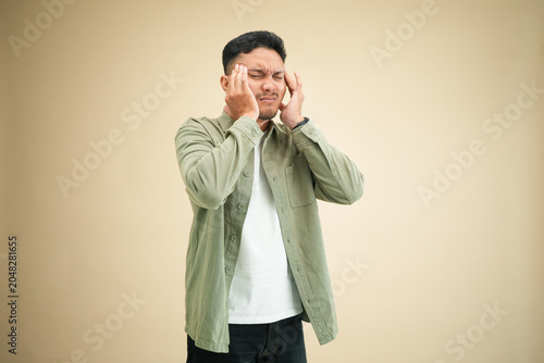 Young Asian man in olive green shirt holding head with both hands in severe distress reflecting mental collapse from financial and economic pressure.
