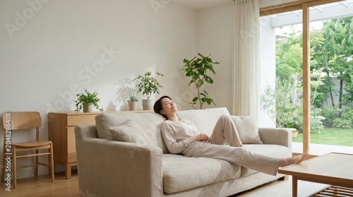 A happy Asian woman relaxing on a sofa features a bright minimalist living room garden background during afternoon sunlight