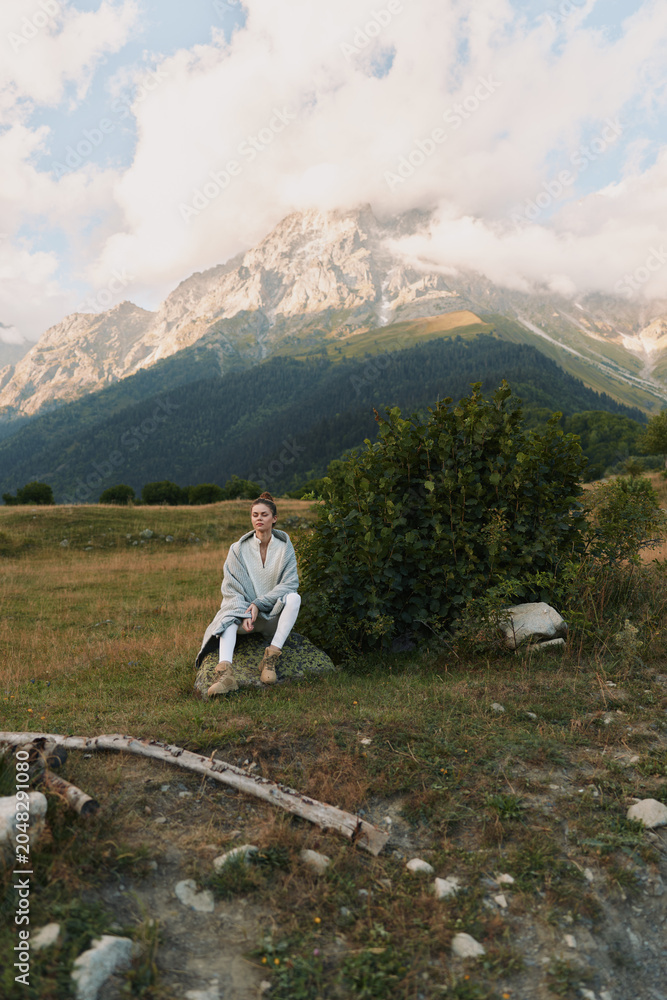 Naklejka premium Quiet outdoor scene with a person sitting on a rock in a grassy field, mountain range in the distance, clear sky, relaxed pose, natural colors, serene lifestyle moment