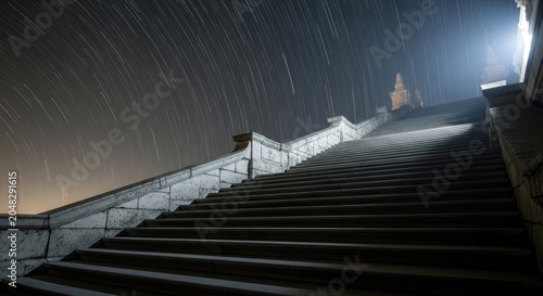 Grand stone staircase leading up to a bright light under a starry night sky.