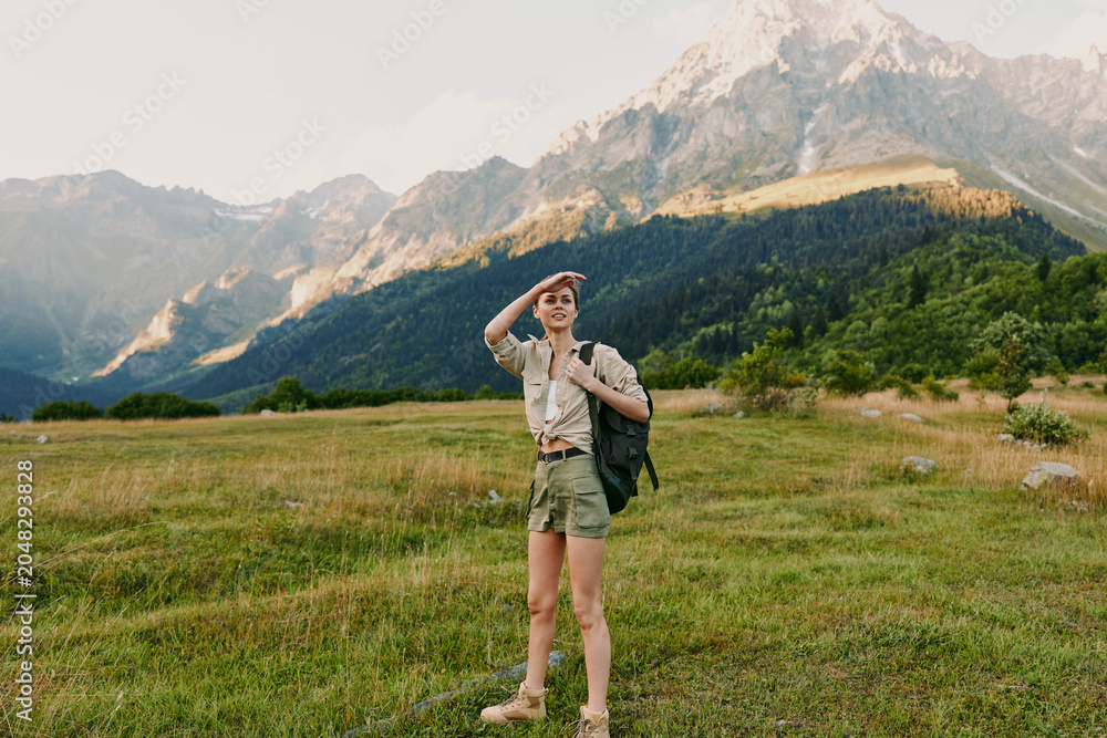 Obraz premium Young woman hiking in a bright meadow beside majestic mountains, carrying a backpack, casual outdoor attire, capturing a spontaneous travel moment in nature