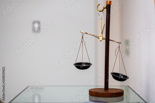 Obraz na plátně Vintage balance scale with black pans hanging on a dark wooden stand, placed on a glass tabletop against a bright blurred wall background, shallow depth of field, soft lighting