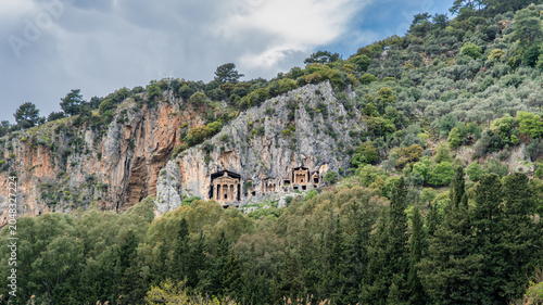Ancient Lycian rock tombs carved into high cliffs surrounded by lush green trees in Dalyan, Turkey. Historic archaeological landmark and heritage scenery.