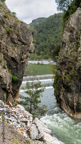Turquoise waters of the Namnam River cascade over a stepped weir between moss covered rocky cliffs and dense pine forest in Mugla, Turkey. Vertical photo