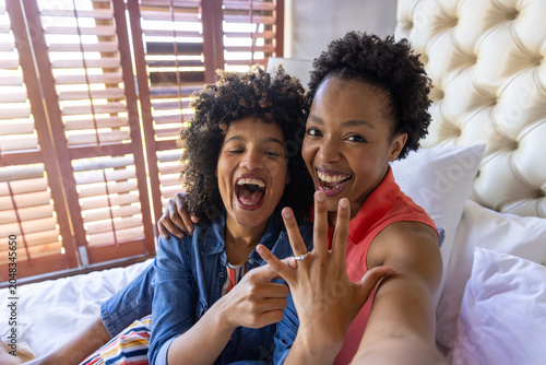 Fotografie Diverse female midadult couple holding phone showing engagement ring on bed in d