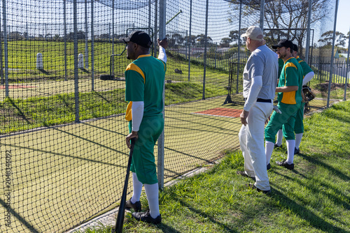 Coach signaling start, batter leaning on net holding bat, teammates watching preparing for practice