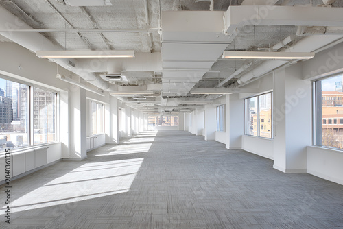 An empty, brightly lit office space with large windows and a view of urban buildings from above. Concrete ceiling and carpeted floor