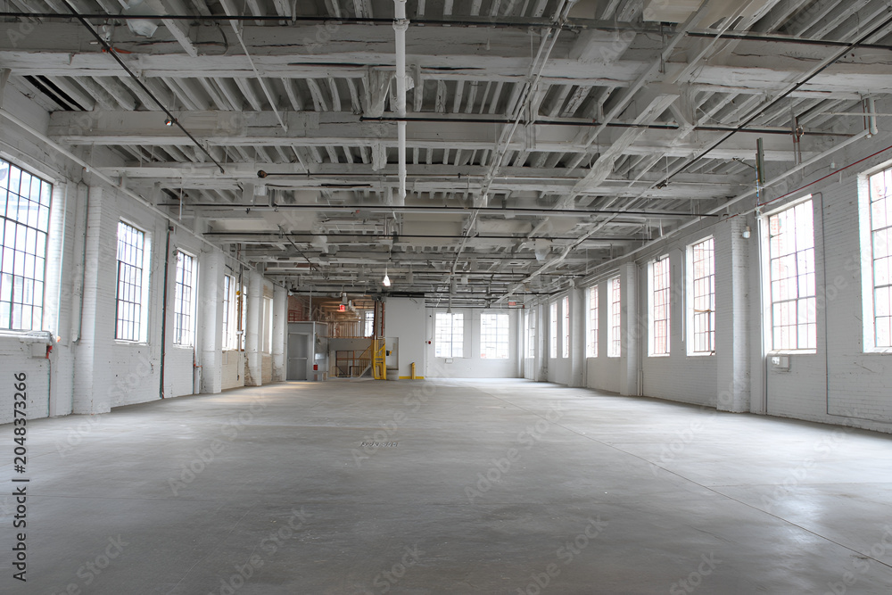 Obraz premium Large, empty, bright interior space with high ceilings, exposed beams, numerous windows, and a smooth concrete floor. The room is light