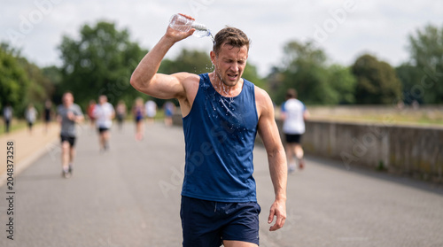 A male runner cools down by pouring water over his head after a strenuous run on a hot day, with other runners in the background.
