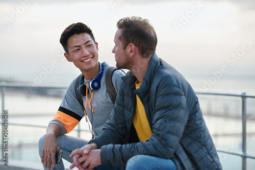 Foto Men, friends and chat on promenade at beach with smile, bonding and conversation on seaside vacation