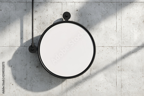 Overhead shot of a blank circular sign with a black frame, mounted on a textured, light-colored concrete wall, casting shadows