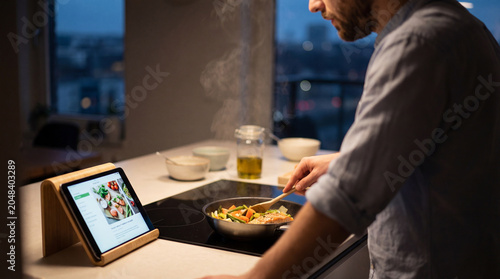 Bearded man cooks healthy dinner in evening kitchen while reading tablet recipe fresh vegetables steam rising from pan smart home wellness meal prep lifestyle content planning daily routine