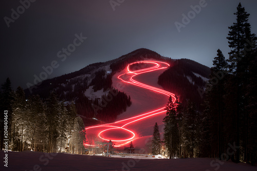 Long exposure shot of a mountain ski slope lit up with red light trails at night. Trees frame the landscape