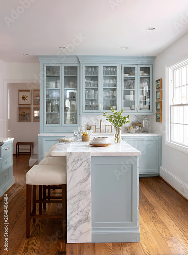 Airy kitchen boasts pale blue cabinetry, marble countertops, an island with seating, and hardwood floors. Framed artwork decorates the walls