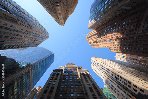 A worm's-eye view, showcasing towering buildings against a clear blue sky. Various architectural styles, textures. Sunlit facades dominate