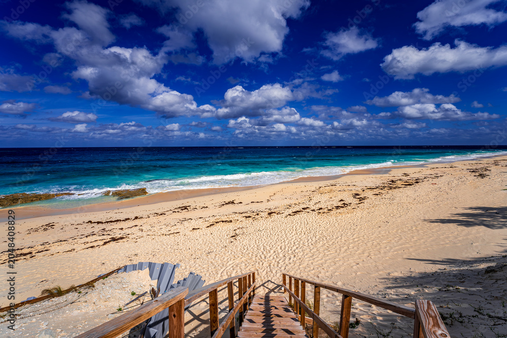 Fototapeta premium Wooden staircase leading down to a pristine sandy beach on Great Guana Cay, Bahamas, with turquoise Caribbean waters, gentle surf, and dramatic cloud formations under a vibrant blue tropical sky.