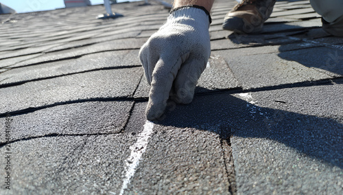 Close-up of a hand in a glove pointing at the seam of a weathered asphalt shingle roof, assessing a potential leak
