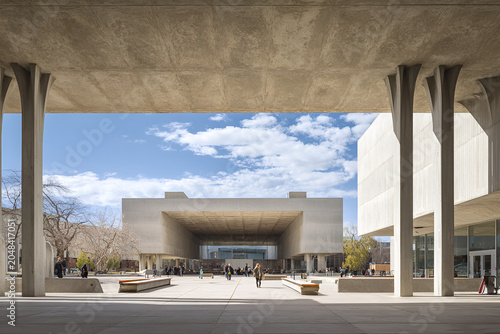 Architectural photograph shows a wide, open-air plaza framed by imposing concrete structures under a bright blue sky