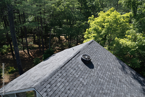 An aerial shot showcases a house roof composed of gray shingles, with a vent. Lush green trees surround the structure