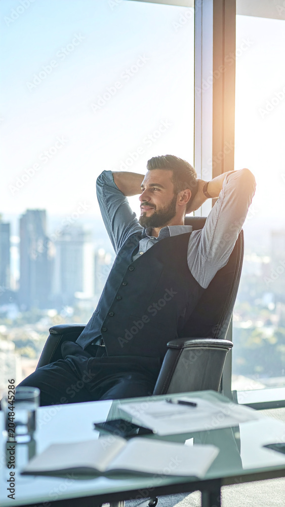 Fototapeta premium Successful businessman relaxes in a highrise office, looking out at a sunny city skyline. Ideal for themes of leadership, career success, wealth, and executive worklife balance.