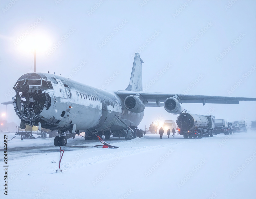 Fototapeta premium A damaged heavy cargo airplane rests on a frozen runway during an extreme arctic blizzard, surrounded by ground service vehicles. Perfect for polar aviation and survival concepts.