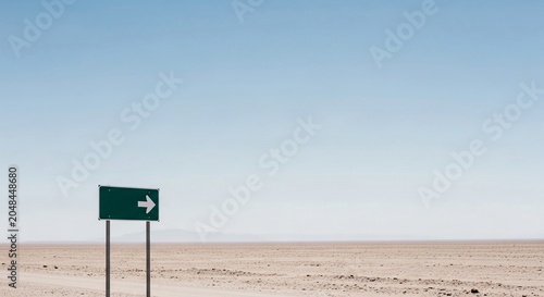 Green directional arrow sign pointing right in a vast desert landscape under a clear blue sky