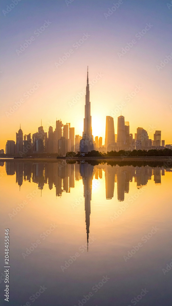 Obraz premium A breathtaking golden sunrise glows behind a modern metropolitan skyline, with an iconic towering skyscraper perfectly reflected in the calm waters. Majestic, serene, and inspiring.
