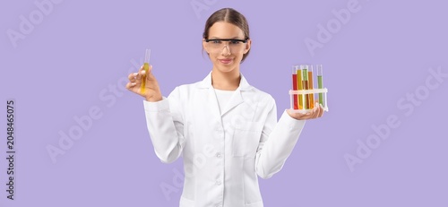 Young female laboratory worker with test tubes on lilac background