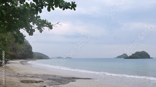 A serene tropical beach view framed by green leaves, showcasing calm blue sea, white sand, and distant islands under a cloudy sky.