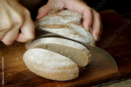 Hands slicing gluten free loaf of brown bread on wooden cutting board. Country bread, homemade pastry.