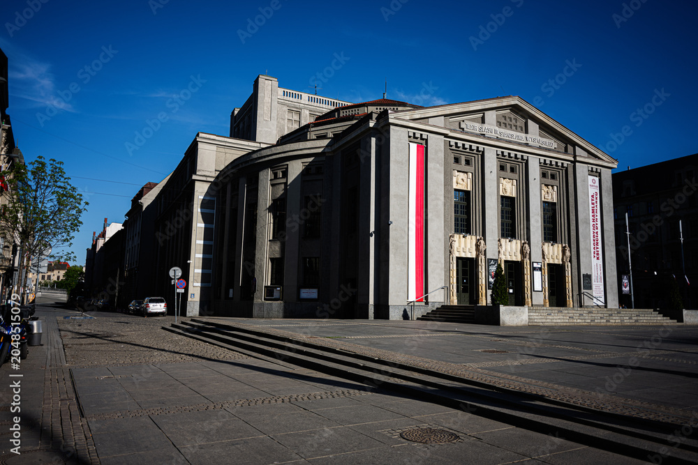 Fototapeta premium City theater building in Katowice Poland with neoclassical facade visitors on steps and quiet urban square under a deep blue sky