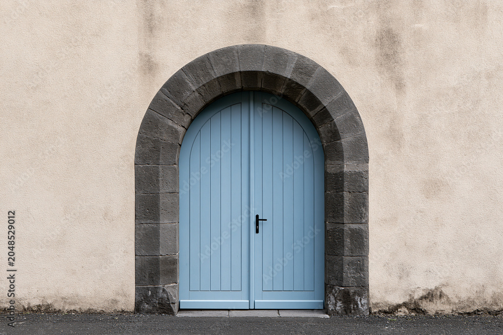 Fototapeta premium Elegant light blue arched double door set within a dark grey stone archway on a textured beige wall, featuring a minimalist and symmetrical architectural design in a clean outdoor setting.