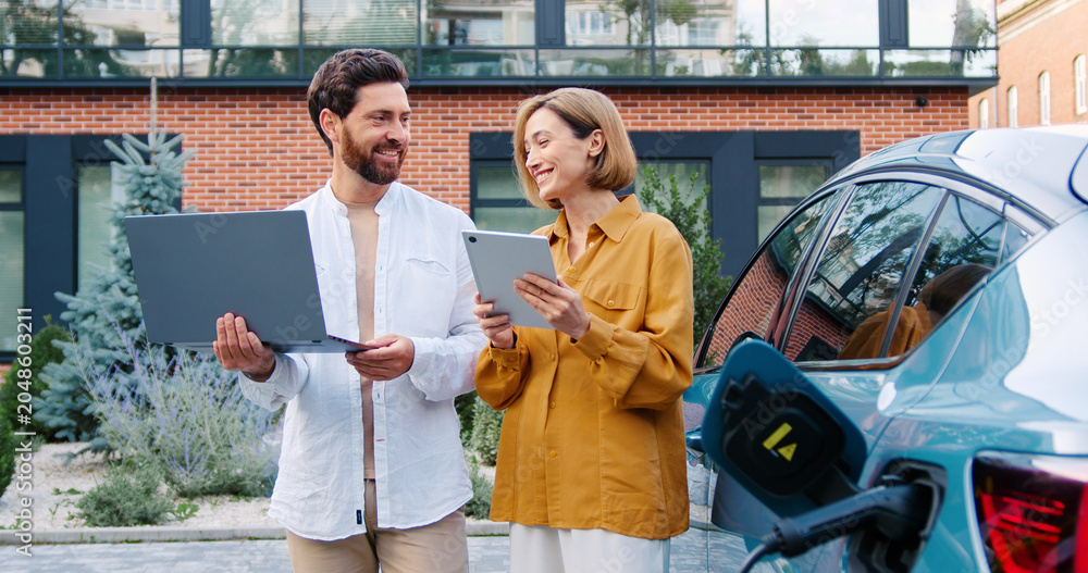 Obraz premium Business man holding a laptop and business woman holding a tablet standing outdoors near a charging electric car, looking at device screens and talking.