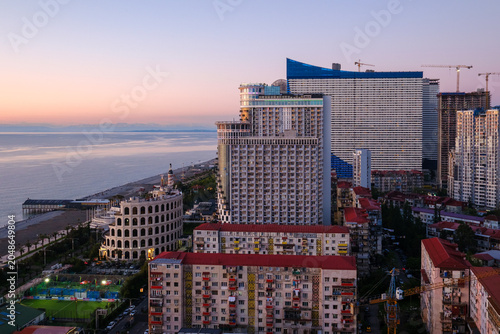 Foto Horizontal landscape photograph showcasing Batumi's modern high-rise architecture at twilight, featuring a soft pink and purple sunset afterglow over the Black Sea with initial city lights turning on